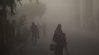 L'immagine mostra una scena di una strada avvolta nella nebbia o nella polvere. In primo piano, una donna con un velo scuro tiene la mano di un bambino, mentre camminano insieme. Sullo sfondo, si possono vedere altre persone che camminano, ma sono poco visibili a causa della foschia. Gli alberi e alcune piante ai lati della strada creano un'atmosfera un po' opprimente. La luce sembra attenuata, dando un'impressione di desolazione.