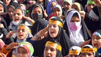تظهر الصورة مجموعة من الفتيات يرتدين الحجاب، وهن يحتفلن أو يشاركن في فعالية جماعية. الفتيات متمركزات في مجموعة، ويبدين حماسًا ونشاطًا، حيث يرفعن أياديهن ويعبرن عن الفرح أو الحماسة. بعضهن يرتدين عصبات على الرأس مكتوب عليها كلمات تبدو تعبيرية. الجو العام في الصورة يوحي بالاحتفال والطاقة الإيجابية.