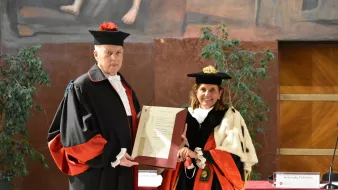 L'immagine mostra due persone vestite in abiti accademici formali, tipici per cerimonie di laurea o conferimenti di titoli. Il soggetto a sinistra indossa un mantello nero con dettagli rossi e un cappello, mentre a destra c'è una donna con un abito simile, ma con un mantello e un cappello che contengono diversi elementi decorativi. Entrambi stanno tenendo un documento o un certificato che sembra essere di rilevanza accademica o ufficiale. Sullo sfondo, ci sono piante e un muro decorato, indicando un ambiente formale e celebrativo.