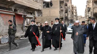 The image shows a group of religious figures walking through a war-torn street. They are dressed in traditional clergy attire, including black robes and crosses, and some are holding staffs. The surroundings are heavily damaged, with buildings showing signs of destruction and debris on the street. In the background, there are a few people and vehicles, indicating a somber atmosphere in a once-bustling area. The overall scene reflects the impact of conflict on the community and highlights the presence of religious leadership amidst the destruction.