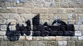 L'immagine mostra un murale su un muro di pietra. Il disegno rappresenta una silhouette di un veicolo, probabilmente un camion, realizzato con vernice nera. Le linee e le forme sono stilizzate, creando un contrasto con la texture della parete di pietra. Sul muro si notano anche resti di manifesti strappati e segni di usura, che aggiungono carattere e storia all'ambiente.
