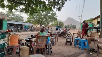 L'image montre une scène de la vie quotidienne dans un village. On y voit plusieurs chaises de différents styles et couleurs, dont certaines sont empilées. À l'arrière-plan, il y a des maisons, des collines, et plusieurs motos. La végétation est présente avec quelques arbres, offrant de l'ombre. On aperçoit également des personnes assises et se déplaçant, ce qui donne un aperçu de l'ambiance locale. Les routes sont poussiéreuses, suggérant une atmosphère rurale.
