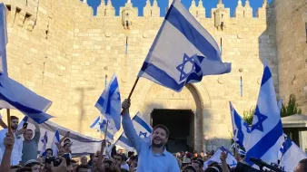 تصویر نشان‌دهنده جمعیتی است که با پرچم‌های اسرائیل در حال جشن و شادی هستند. در میان این جمعیت، فردی در حال بلند کردن پرچم و ابراز شادی است. پس‌زمینه تصویر یک دروازه تاریخی و دیوار سنگی نمایان است که نشان‌دهنده یک مکان مهم است. افراد حاضر در تصویر لباس‌های مختلفی به تن دارند و فضای کلی عکس نشان‌دهنده شور و نشاط است.
