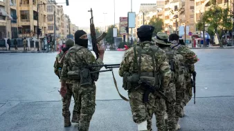 L'image montre un groupe de soldats armés marchant dans une rue urbaine. Ils portent des uniformes militaires et sont équipés d'armes. L'environnement semble urbain, avec des bâtiments en arrière-plan. L'ambiance est sérieuse, suggérant une situation potentiellement tendue. Les soldats portent également des masques, ce qui peut indiquer des préoccupations de sécurité.