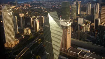 L'image montre une vue aérienne d'une métropole moderne, probablement une grande ville avec des gratte-ciels. On observe des bâtiments de différents styles architecturaux, certains avec des façades en verre réfléchissant. Au centre, un bâtiment en forme de pyramide avec des bords angulaires se détache, portant le mot "BANK". La ville est entourée de collines et d'un ciel partiellement nuageux, ce qui indique une atmosphère dynamique. Les routes serpentent à travers les bâtiments, suggérant une activité urbaine animée.