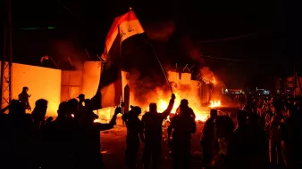 L'image montre une scène nocturne animée, probablement lors d'une manifestation ou d'une émeute. Des personnes brandissent un drapeau, en l'occurrence le drapeau irakien, tandis que des flammes s'élevées en arrière-plan, suggérant des tensions ou des troubles. Les silhouettes des manifestants sont bien visibles, et l'ambiance est chargée d'énergie et d'émotion. La lumière des feux crée un contraste fort avec l'obscurité de la nuit.