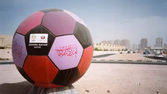 L'immagine mostra un'enorme palla da calcio decorata, posizionata in un'area all'aperto. La palla presenta colori vivaci, inclusi viola e nero, e ha delle scritte decorative, probabilmente in arabo. Sul fronte, si vede un cartello con la scritta "BIDDING NATION" e il nome "QATAR", che indica il ruolo del Qatar come nazione candidata per un evento sportivo. Sullo sfondo, si intravedono edifici moderni e strade, in un contesto urbano. L'atmosfera è illuminata da una luce chiara, suggerendo una giornata di sole.