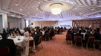 L'image montre une salle de conférence ou de réunion spacieuse, avec de nombreuses personnes assises autour de tables rondes. Les participants semblent engagés dans une discussion ou une présentation, avec un panel sur une estrade à l'avant. La décoration est moderne, avec un plafond orné et un éclairage doux. Les personnes sont habillées de manière formelle, ce qui indique un événement professionnel ou officiel.