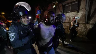 تظهر الصورة مجموعة من رجال الشرطة الذين يقومون باعتقال رجل. يبدو الرجل عاري الصدر ويظهر عليه علامات التوتر أو المقاومة. تحيط به عناصر من الشرطة يرتدون معدات حماية، ويبدو أنهم يتخذون إجراءات للسيطرة عليه. الخلفية توحي بأن المكان ربما كان شاهداً على مظاهر من التوتر أو الاضطراب، مع وجود إضاءة خافتة تشكل جوًا متوترًا.