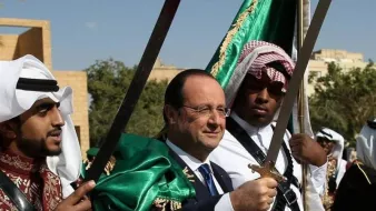 تظهر في الصورة مجموعة من الأشخاص يرتدون أزياء تقليدية. في المنتصف، يقف رجل يرتدي بدلة رسمية مع ابتسامة، محاطًا بشخصين آخرين يحمل كل منهما سيفًا. يزخر المشهد بالألوان، حيث يرتدي الشخصان الآخران ألبسة تقليدية بألوان بارزة، مع بعض الزينة الثقافية. الخلفية تضم أشجار وبعض المباني، وتبدو السماء صافية.