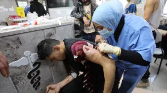 L'immagine mostra una scena in un ambiente sanitario. Un giovane uomo, visibilmente ferito, è seduto su una sedia mentre una professionista sanitaria, vestita con una divisa blu e con guanti, sta medicando una sua ferita sul braccio. Il suo volto mostra preoccupazione, e l'atmosfera appare tesa. Sullo sfondo si possono vedere altre persone e attrezzature mediche, suggerendo un contesto di emergenza.