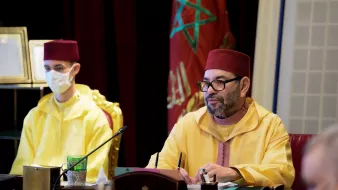 En la imagen se puede ver a dos personas en un entorno formal. A la derecha, un hombre lleva una vestimenta tradicional marroquí de color amarillo y un fez. Este hombre parece estar en una reunión o conferencia. A la izquierda, hay una persona más joven que también lleva una vestimenta amarilla y un fez rojo, además de llevar mascarilla. Ambos están sentados frente a una mesa con documentos y otros elementos, en un ambiente que sugiere un evento importante.