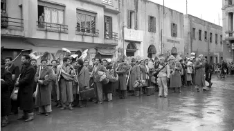 L'image montre une file de personnes alignées dans une rue apparemment urbaine. Les individus portent des vêtements de travail et des sacs, ce qui suggère qu'ils attendent quelque chose, peut-être un transport ou un appel. En arrière-plan, on peut voir des bâtiments avec des fenêtres et des balcons, ainsi que des éléments architecturaux qui indiquent une époque passée. L'atmosphère semble sérieuse, et le pavement est mouillé, peut-être à cause de la pluie.