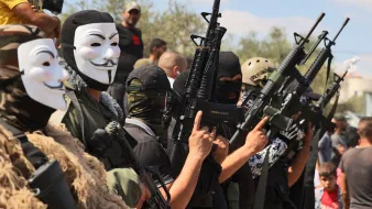 L'image montre un groupe de personnes portant des masques et des vêtements militaires, tenant des fusils. Les visages des individus sont principalement cachés, et ils semblent formés en ligne, chacun montrant une arme. En arrière-plan, on aperçoit d'autres personnes observant la scène. L'atmosphère semble tendue et engagée.
