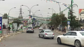 L'image montre une rue bordée de drapeaux et de bannières, probablement en préparation d'un événement ou d'une célébration. On peut voir plusieurs voitures circulant sur la route. À côté de la route, il y a des panneaux avec des affiches, ce qui indique une zone urbaine active. Des personnes se trouvent également sur le trottoir, et des arbres et des lampadaires sont présents le long de la rue, donnant un aspect dynamique à la scène. Le ciel semble nuageux, suggérant une ambiance calme.