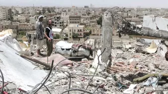تظهر الصورة مشهدًا من مدينة مدمرة، حيث يقف رجلان على أنقاض مبنى. خلفهما، ترتفع مجموعة من المباني في المدينة، وهي تبدو متضررة بشكل كبير. يبدو أن البيئة المحيطة تعكس آثار الدمار، مع الحطام والمخلفات المنتشرة في كل مكان. السماء تبدو مشمسة، مما يبرز الف contrast بين الدمار والبيئة المحيطة.