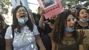 L'immagine mostra un gruppo di persone che partecipa a una manifestazione. Alcuni manifestanti tengono in mano cartelli con fotografie e slogan, mentre gridano in segno di protesta. Le persone indossano mascherine e sembrano essere molto coinvolte emotivamente, esprimendo le loro richieste con forza. Il contesto sembra essere urbano, con alberi e edifici sullo sfondo.