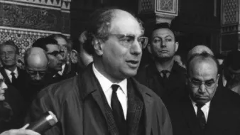 L'image montre un homme en train de prendre la parole devant un groupe de personnes. Il porte un manteau et des lunettes. Derrière lui, il y a plusieurs hommes et femmes qui l'écoutent attentivement. L'environnement semble formel, et il y a une atmosphère de rassemblement ou de cérémonie. En arrière-plan, on aperçoit des éléments d'architecture qui suggèrent un lieu historique ou important.