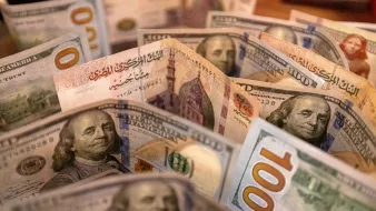 The image features a collection of banknotes, specifically U.S. hundred-dollar bills. The bills are arranged in a way that showcases their distinctive green and blue colors, with the face of Benjamin Franklin prominently displayed on the front. Additionally, some bills feature Arabic script, indicating a mix with other currencies or designs. The background is slightly blurred, focusing attention on the detailed designs of the banknotes.