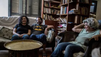 L'immagine mostra un ambiente domestico accogliente, con un soggiorno arredato in modo informale. Ci sono tre persone sedute su poltrone e divani. Due di loro sorridono, mentre un'altra donna, con capelli grigi e ricci, appare rilassata. In mezzo c'è un tavolino rotondo di metallo. Sullo sfondo, ci sono librerie piene di libri, che conferiscono un'atmosfera calda e familiare all'ambiente. Sul pavimento, si intravede un cane.
