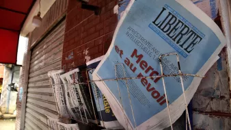 L'image montre un présentoir de journaux, où plusieurs titres sont visibles. Le journal en premier plan affiche le mot "LIBERTÉ" en lettres majuscules, accompagnés d'un sous-titre qui semble évoquer un sujet d'actualité. Les autres journaux à côté sont disposés de manière ordonnée, avec des titres variés, ce qui suggère un point de vente de presse dans un environnement urbain. Le mur en arrière-plan, ainsi que le toît rouge au-dessus, ajoutent à l'atmosphère locale de la scène.