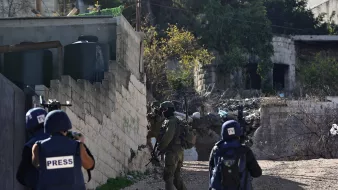 En la imagen se pueden ver varios individuos. A la izquierda, hay personas vestidas con chalecos que tienen la palabra "PRESS" correspondiente a la prensa o periodistas. A la derecha, hay un soldado con equipo militar que sostiene un arma y parece estar en una postura de vigilancia. El entorno muestra un área urbana con edificios y escombros, lo que sugiere un contexto de tensión o conflicto. La situación parece ser de cobertura de un evento relacionado con la seguridad o una operación militar.