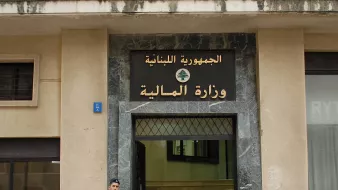 L'image montre un bâtiment avec une façade en pierre, portant une inscription en arabe et en français. On y voit un soldat en uniforme, armé, qui se tient devant l'entrée du bâtiment. Le soldat semble en position de garde, et à côté de lui, on peut remarquer des éléments architecturaux typiques des bâtiments gouvernementaux. L'inscription indique que l'édifice est le ministère des Finances du Liban.
