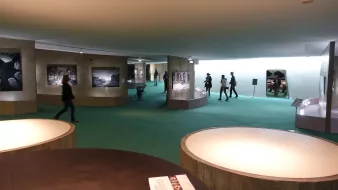 L'image présente un espace d'exposition, probablement dans un musée. On peut y voir des murs de couleur claire avec des photographies accrochées. Le sol est recouvert d'un tapis vert, créant une atmosphère accueillante. Des visiteurs se déplacent dans cet espace, explorant les œuvres exposées. Au premier plan, il y a plusieurs tables ou surfaces circulaires, apparemment utilisées pour des brochures ou d'autres informations. L'éclairage est doux et met en valeur les œuvres sur les murs.