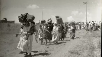 L'image montre un groupe de personnes marchant le long d'un chemin poussiéreux. Certaines femmes portent de lourdes charges sur la tête, tandis que d'autres tiennent des enfants par la main. La scène semble illustrer un moment de déplacement ou de migration, avec des paysages arides en arrière-plan. Des nuages blancs ponctuent le ciel, et on peut voir des lignes électriques au loin. L'atmosphère de la photo évoque la lutte et la détermination des personnes en marche.