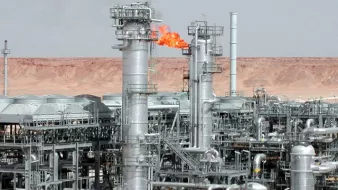L'image montre une installation industrielle, probablement liée à l'industrie pétrolière ou gazière. On peut voir plusieurs tours métalliques, dont certaines émettent des flammes, indiquant une combustion, peut-être de gaz résiduel. L'arrière-plan présente un paysage désertique, caractérisé par des collines ou des montagnes sèches, typique des zones où se trouvent des installations énergétiques. Les structures sont en métal et semblent sophistiquées, témoignant d'une technologie avancée utilisée dans cette industrie.