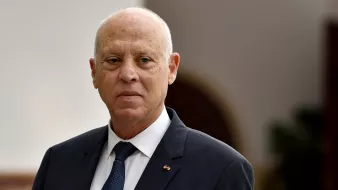 The image shows a man dressed in a suit with a tie, standing in what appears to be an official or dignified environment. He has a serious expression on his face and is looking slightly away from the camera. The background is softly blurred, suggesting it is a formal setting.
