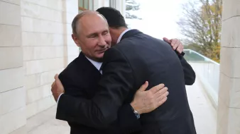الصورة تظهر شخصين يتعانقان في مشهد يبدو وديًا. الشخص الأول يرتدي بدلة سوداء وقميصًا أبيض، بينما الشخص الثاني يرتدي بدلة داكنة أيضًا. الخلفية تظهر جزءًا من مبنى وأشجار، مما يوحي بأن اللقاء يحدث في مكان رسمي أو خارجي. تعبر تعابير الوجه عن اللحظة العاطفية لهذا اللقاء.