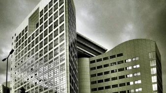 L'image montre un bâtiment moderne avec une architecture contemporaine. La structure est composée de plusieurs angles et surfaces réfléchissantes, donnant une impression de sophistication. Les vitres sont grandes, et le design présente des lignes épurées. Le ciel en arrière-plan est nuageux, ce qui peut créer une ambiance dramatique. Les tons de l'image sont en noir et blanc, accentuant les contrastes entre la lumière et l'ombre sur la façade du bâtiment.