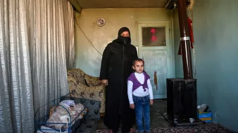 L'image montre une femme portant une tenue noire et un voile, se tenant à côté d'une petite fille. Elles sont à l'intérieur d'une pièce qui semble modeste, avec des murs verts pâles et un tapis au sol. À gauche, il y a une couchette avec un bébé endormi. Un poêle en métal est visible dans un coin de la pièce, et des rideaux en tissu léger sont tirés. L'ambiance est familiale, mais la simplicité du décor reflète une certaine précarité.
