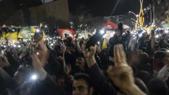 در این تصویر، جمعیتی پرشور و هیجان‌زده دیده می‌شود که در یک شب تاریک گرد هم آمده‌اند. افراد حاضر در مراسم، پرچم‌هایی با رنگ‌های مختلف را در دست دارند و برخی از آن‌ها نیز با استفاده از گوشی‌های خود چراغ‌ قوه‌ها را روشن کرده‌اند. فضای عمومی شلوغ و مملو از شور و احساسات اجتماعی یا سیاسی به نظر می‌رسد. جماعت به سمت دوربین یا نقطه‌ای خاص معطوف شده و برخی با دست خود علامت‌هایی را نشان می‌دهند.