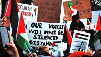 L'immagine mostra una folla di persone impegnate in una manifestazione. La maggior parte dei manifestanti tiene in mano cartelli con diverse scritte e bandiere, molte delle quali sembrano rappresentare il Palestinese. Tra i cartelli, uno evidenzia il messaggio "Our voices will never be silenced," che esprime determinazione e unità. Atmosfera di protesta e attivismo è palpabile, con la presenza di simboli politici e sociali.