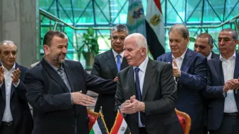 L'image montre un groupe de personnes lors d'une cérémonie officielle. À l'avant, deux hommes se serrent la main avec des sourires, suggérant un accord ou un partenariat. Ils sont entourés d'autres individus qui applaudissent, indiquant un moment de célébration. À l'arrière-plan, on peut distinguer des drapeaux, probablement ceux de la Palestine et de l'Égypte, ce qui laisse penser que cette rencontre est d'une importance politique. L'environnement semble être un bâtiment officiel, avec de grandes fenêtres laissant entrer la lumière.
