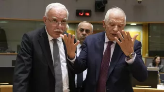 L'image montre deux hommes en discussion animée, debout dans un cadre de réunion. L'un d'eux a des cheveux gris et porte des lunettes, tandis que l'autre est également âgé avec des cheveux clairs. Ils semblent gesticuler avec leurs mains pour souligner leurs propos. En arrière-plan, une autre personne les observe. L'environnement paraît être une salle de conférence, avec des tables et des collègues travaillant.