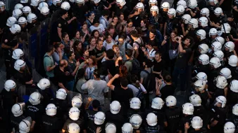 L'image montre une scène de manifestation où une foule de personnes se trouve face à des policiers en tenue anti-émeute. Les manifestants semblent discuter ou protester, tandis que les policiers forment une ligne serrée. L'atmosphère est tendue, et une partie de la foule semble concentrée sur un point, indiquant une interaction ou un affrontement verbal. Les casques des policiers et leur équipement renforcent l'impression de confrontation.