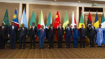 L'image montre un groupe de personnes debout en rangée, saluant. Elles se trouvent devant une série de drapeaux représentant différents pays. Les personnes portent des costumes formels et semblent participer à un événement diplomatique ou à un sommet international. Les drapeaux en arrière-plan incluent ceux de l'Union africaine, de la Chine, ainsi que d'autres nations. L'ambiance générale semble conviviale et respectueuse.