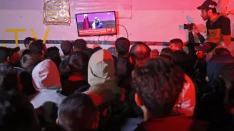 L'immagine mostra un gruppo di persone affollate intorno a un televisore. Sullo schermo è visibile un uomo che parla, mentre la parte inferiore dello schermo mostra un testo in arabo. L'illuminazione dell'ambiente è scarsa e i partecipanti indossano vari tipi di abbigliamento, inclusi cappucci e giacche. Alcuni presenti sono distratti dal loro smartphone o sembrano concentrati su ciò che viene trasmesso in TV.