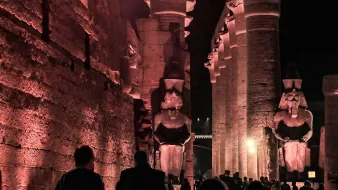 L'image présente un site historique, probablement des ruines égyptiennes, éclairées par des lumières tamisées, ce qui crée une atmosphère mystérieuse. On peut voir de grandes statues de pharaons ou de divinités, flanquant une structure impressionnante faite de colonnes. Les visiteurs se déplacent à travers le site, ajoutant une dimension humaine à cette scène ancienne, où l'architecture et les sculptures témoignent de la grandeur de la civilisation égyptienne. Les ombres et la lumière jouent un rôle important, accentuant les détails des sculptures et des murs.