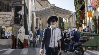 L'image montre un homme marchant dans une rue, portant un costume noir et un chapeau. Il a un masque facial et est vêtu d'un tefillin sur son bras. En arrière-plan, on peut voir un endroit semblant être un lieu de prière, avec des gens habillés en vêtements traditionnels. Des panneaux et de la végétation décorent le paysage urbain. La scène évoque une atmosphère communautaire et religieuse.