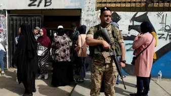 La imagen muestra a un soldado de pie con un rifle, vigilando una entrada. Al fondo, hay un grupo de mujeres, algunas de las cuales llevan hijab, esperando. La escena parece tener lugar en un ambiente urbano, con murales o grafitis en las paredes cercanas. El soldado parece atento y en posición de guardia, mientras las mujeres se agrupan en la entrada.