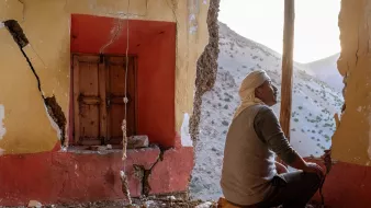 L'image montre un homme assis dans une pièce aux murs endommagés, avec des fissures visibles. À sa gauche, on aperçoit une fenêtre en bois, et à droite, le mur est abîmé, laissant entrevoir l'extérieur. La lumière naturelle pénètre dans la pièce, créant une ambiance mélancolique. L'homme semble contemplatif, perdu dans ses pensées, dans un décor qui suggère l'oubli et la dégradation.
