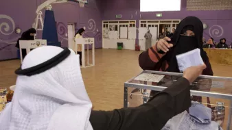 L'image montre une scène d'élection dans un environnement intérieur. Au premier plan, un homme portant un kandora et un ghutrah se tient derrière une urne électorale. Il regarde une femme voilée qui tend un bulletin de vote, le visage partiellement caché par un niqab. En arrière-plan, on peut voir d'autres personnes et des isoloirs, indiquant que le processus de vote est en cours dans cet espace, qui semble être une salle de sport ou un centre communautaire. La décoration du mur est colorée, ajoutant une atmosphère dynamique à l'événement.
