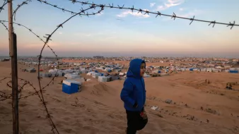 Nell'immagine si vede un bambino che cammina su una duna di sabbia. Indossa una giacca blu e appare felice mentre si muove. Sullo sfondo, un campo profughi si estende all'orizzonte, con molte tende e strutture temporanee. Una recinzione di filo spinato si trova in primo piano, evidenziando il contesto difficile in cui il bambino e gli altri abitanti del campo vivono. Il cielo è sereno, con sfumature di blu e arancio al tramonto.