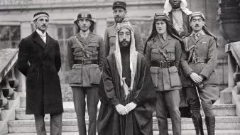 L'image montre un groupe de six hommes posant ensemble. Ils sont vêtus de différentes tenues militaires et traditionnelles. Au centre, un homme en robe traditionnelle, portant une tête couronnée, semble être le principal sujet. À sa gauche, un autre homme en uniforme militaire. Les autres sont également en uniformes variés, indiquant peut-être des origines et des rôles différents. L'arrière-plan montre une architecture qui semble historique. L'ensemble dégage une atmosphère de confluence culturelle et de collaboration.