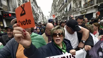 The image depicts a crowd of protesters gathered in an urban setting, likely during a demonstration. A woman in the foreground is holding an orange sign that reads "l'vote" while wearing sunglasses and a green top. The crowd behind her appears to be a mix of men and women, some holding signs and banners. The atmosphere conveys a sense of urgency and solidarity among the protesters, who seem to be advocating for a political cause. The background includes urban architecture, suggesting a city environment.