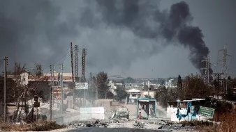 L'image montre une scène de destruction et de conflit. On y voit une route endommagée, avec des débris et des voitures laissées à l'abandon. À l'arrière-plan, un épais nuage de fumée noire s'élève, probablement en raison d'incendies ou d'explosions. Le paysage semble désolé, avec des bâtiments en ruine et une atmosphère de désolation. La présence de poteaux électriques et de panneaux publicitaires ajoute au contexte urbain de l'environnement. L'ensemble de l'image évoque des situations de crise et de guerre.